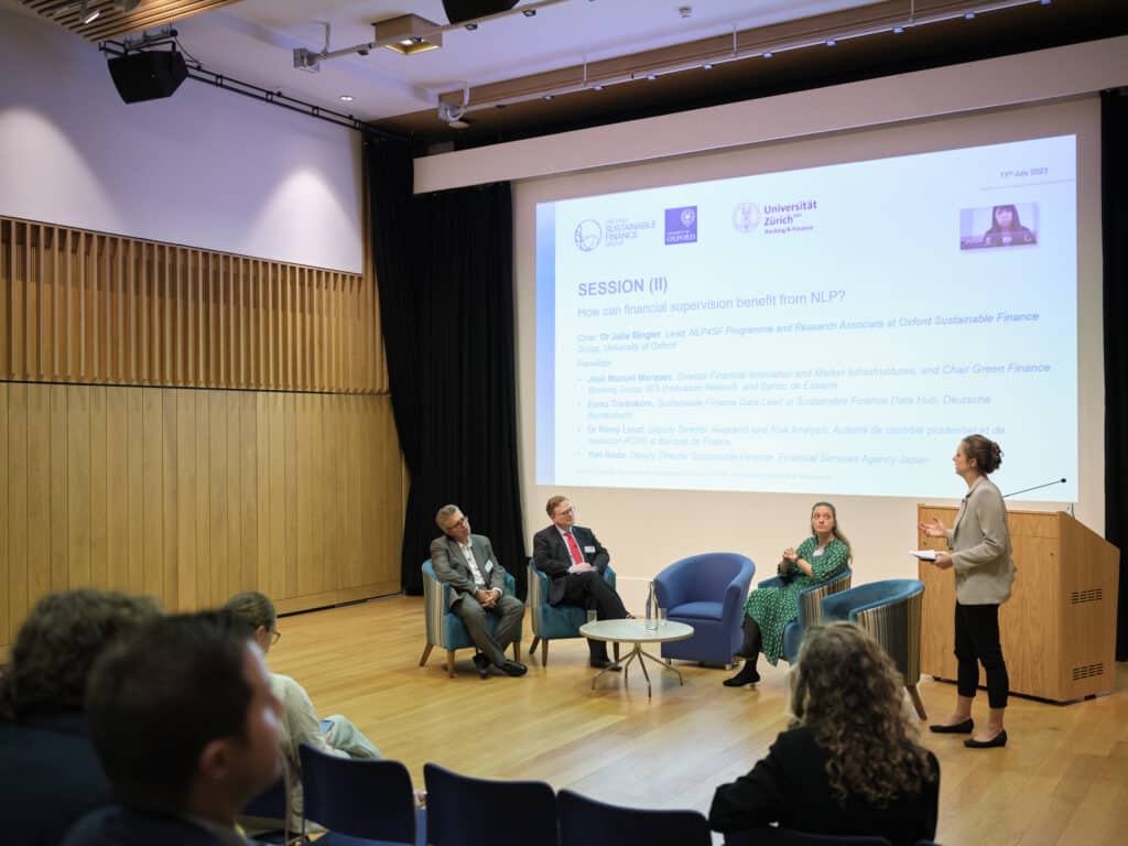 NLP4SF Gallery three people sat on stage looking at woman on stage talking. The screen behind them is banded Oxford Sustainable Finance Group, University of Oxford and Universitat Zurich.